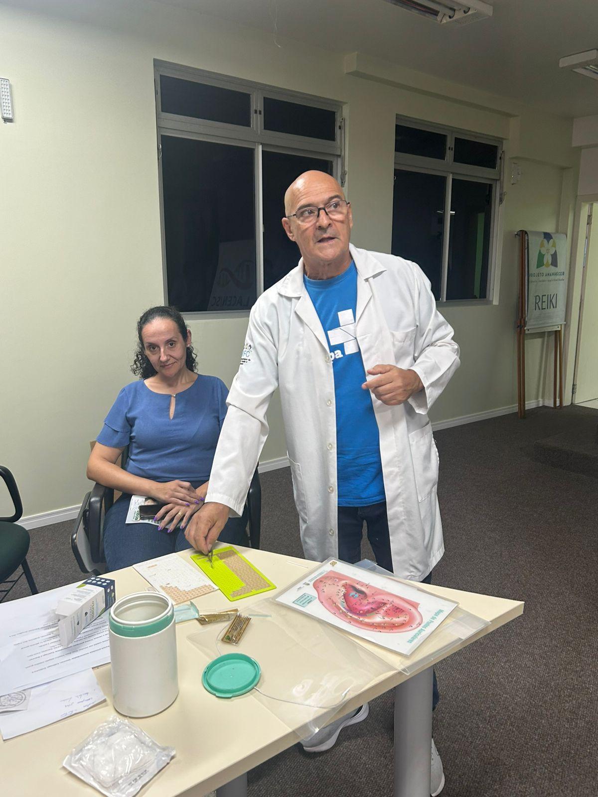 Profissional da saúde apresenta materiais e técnicas de auriculoterapia durante atividade educativa em ambiente interno. Sobre a mesa há cartazes anatômicos da orelha, sementes para aplicação terapêutica e materiais utilizados na prática integrativa. Ao fundo, participante acompanha a apresentação sentada, com placa indicando atividade de reiki no local.