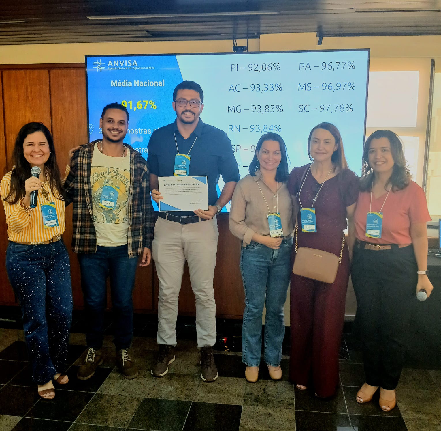 Seis pessoas posam lado a lado em um ambiente interno, em frente a uma tela de apresentação da ANVISA com dados percentuais por estado. No centro, um homem segura um certificado de reconhecimento, enquanto os demais sorriem para a foto. Todos utilizam crachás de identificação. À esquerda, uma mulher segura um microfone. O grupo está em um espaço com parede de madeira e piso quadriculado, indicando um evento ou apresentação oficial.