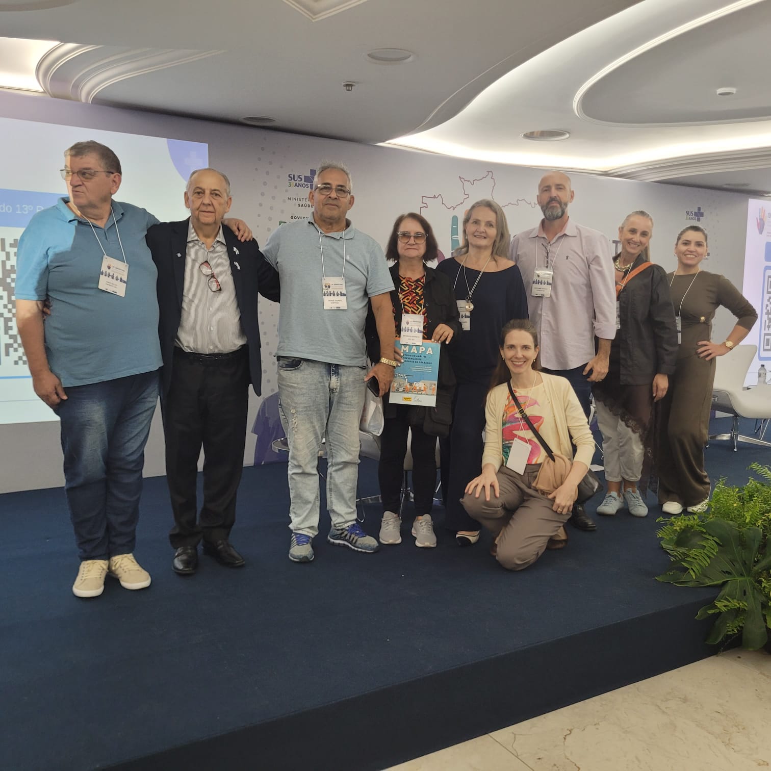 Fotografia de um grupo de dez participantes do 13º RENASTTÃO reunidos sobre um palco durante o evento. Nove pessoas estão em pé lado a lado, usando crachás de identificação, enquanto uma participante está ajoelhada à frente do grupo. Ao fundo, há um painel institucional com referências aos 30 anos do SUS e elementos gráficos relacionados ao evento. Uma das participantes segura um exemplar do documento 'MAPA'. O ambiente é um auditório com iluminação moderna no teto e decoração com plantas na lateral do palco. Todos posam para a foto em um momento de integração e representação da delegação de Santa Catarina.