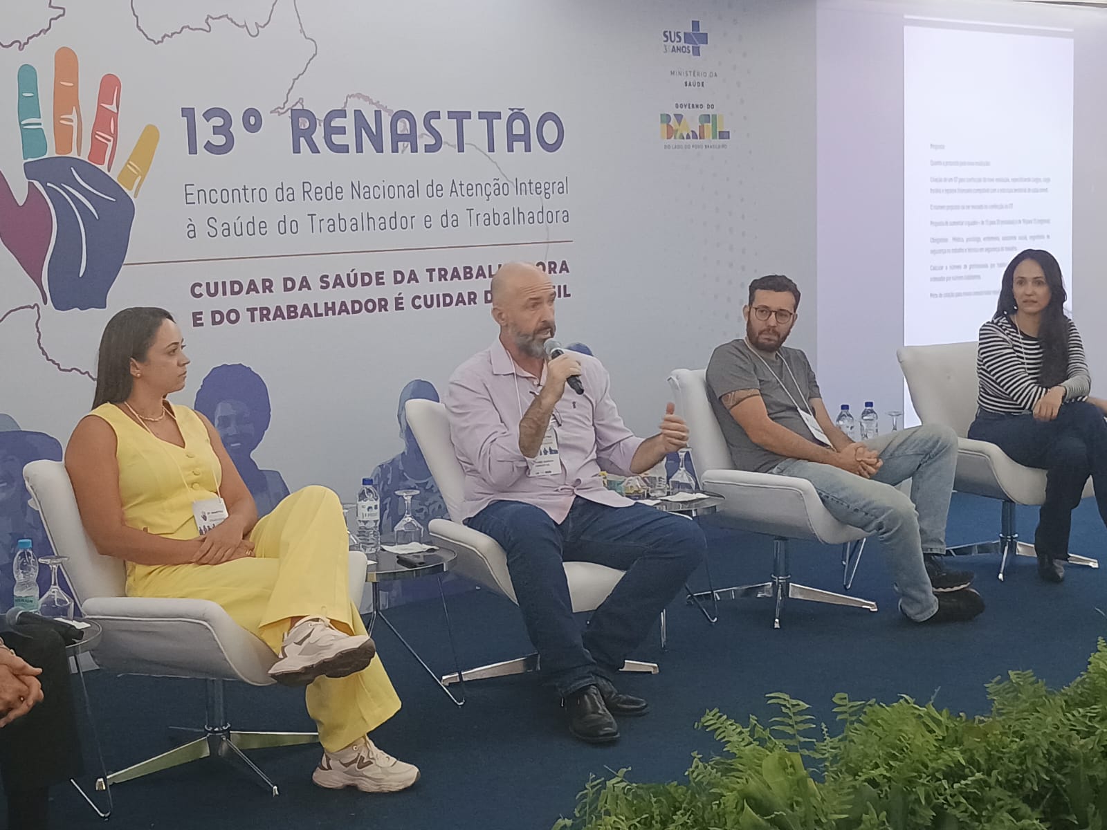 Fotografia de uma mesa de debate durante o 13º RENASTTÃO. Quatro participantes estão sentados em poltronas brancas sobre um palco, enquanto um dos debatedores, ao centro, fala ao microfone e gesticula durante sua apresentação. Ao fundo, um painel exibe a identidade visual do evento com o texto '13º RENASTTÃO – Encontro da Rede Nacional de Atenção Integral à Saúde do Trabalhador e da Trabalhadora' e a mensagem 'Cuidar da saúde da trabalhadora e do trabalhador é cuidar do Brasil'. O ambiente é um auditório com estrutura para palestras e debates, incluindo mesas de apoio, garrafas de água e projeção de conteúdo em tela lateral.