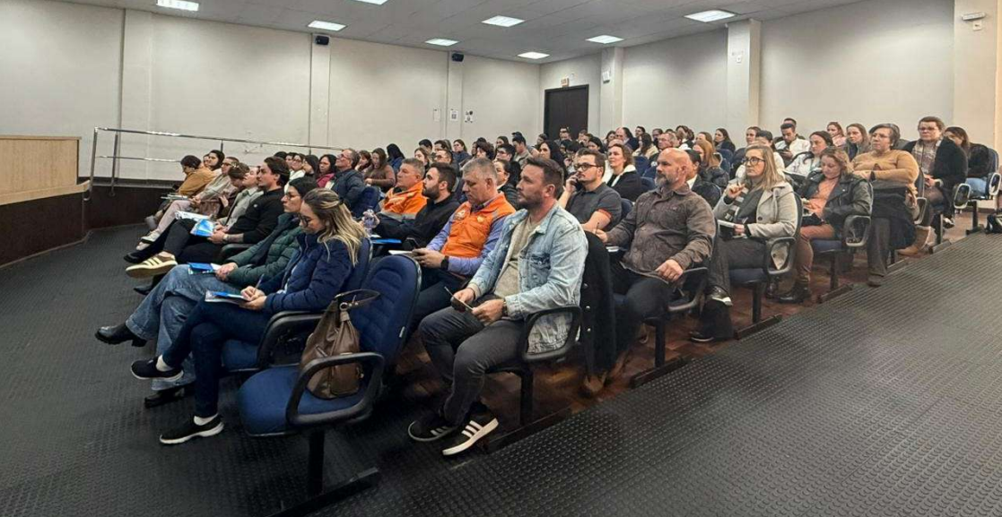 Auditório com dezenas de participantes sentados em cadeiras, acompanhando uma capacitação presencial. O público é composto por profissionais de diferentes áreas, distribuídos em fileiras voltadas para a frente da sala. Alguns fazem anotações enquanto assistem à apresentação. O ambiente é amplo, com iluminação artificial e estrutura adequada para eventos e treinamentos. A imagem registra atividade de qualificação intersetorial sobre a atuação do setor saúde em abrigos temporários.