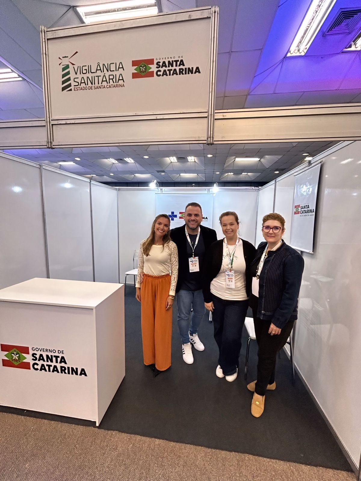 Quatro pessoas posam para foto dentro de um estande da Vigilância Sanitária do Estado de Santa Catarina, com painéis brancos ao redor e logotipos da Vigilância Sanitária e do Governo de Santa Catarina na parte superior e na frente do balcão. O grupo está sorrindo e usando crachás de identificação em um ambiente de evento ou congresso.