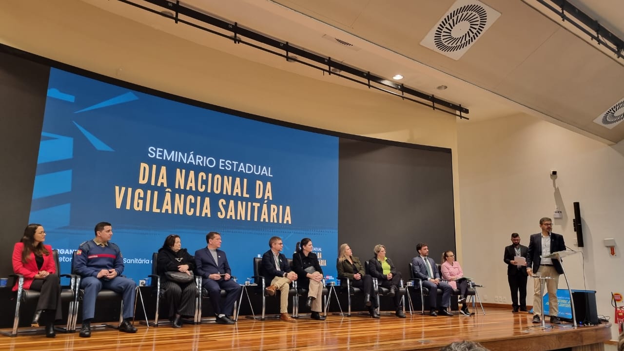 A imagem mostra um auditório durante o Seminário Estadual – Dia Nacional da Vigilância Sanitária. No palco, há uma grande tela de fundo azul com o nome do evento em letras brancas e amarelas. Sentadas em cadeiras pretas, diversas autoridades e convidados compõem a mesa oficial, incluindo homens e mulheres, alguns com trajes formais e outros com uniformes institucionais.  À direita, um homem de terno claro e blazer escuro fala ao microfone em um púlpito, enquanto outras duas pessoas estão em pé ao fundo, uma delas segurando papéis. O ambiente tem iluminação interna quente, piso de madeira e teto claro com sistemas de ventilação visíveis.